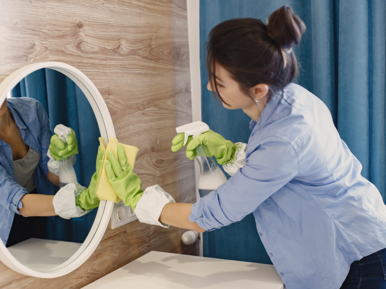 Housewife woking at home. Lady in a blue shirt. Woman clean mirror.